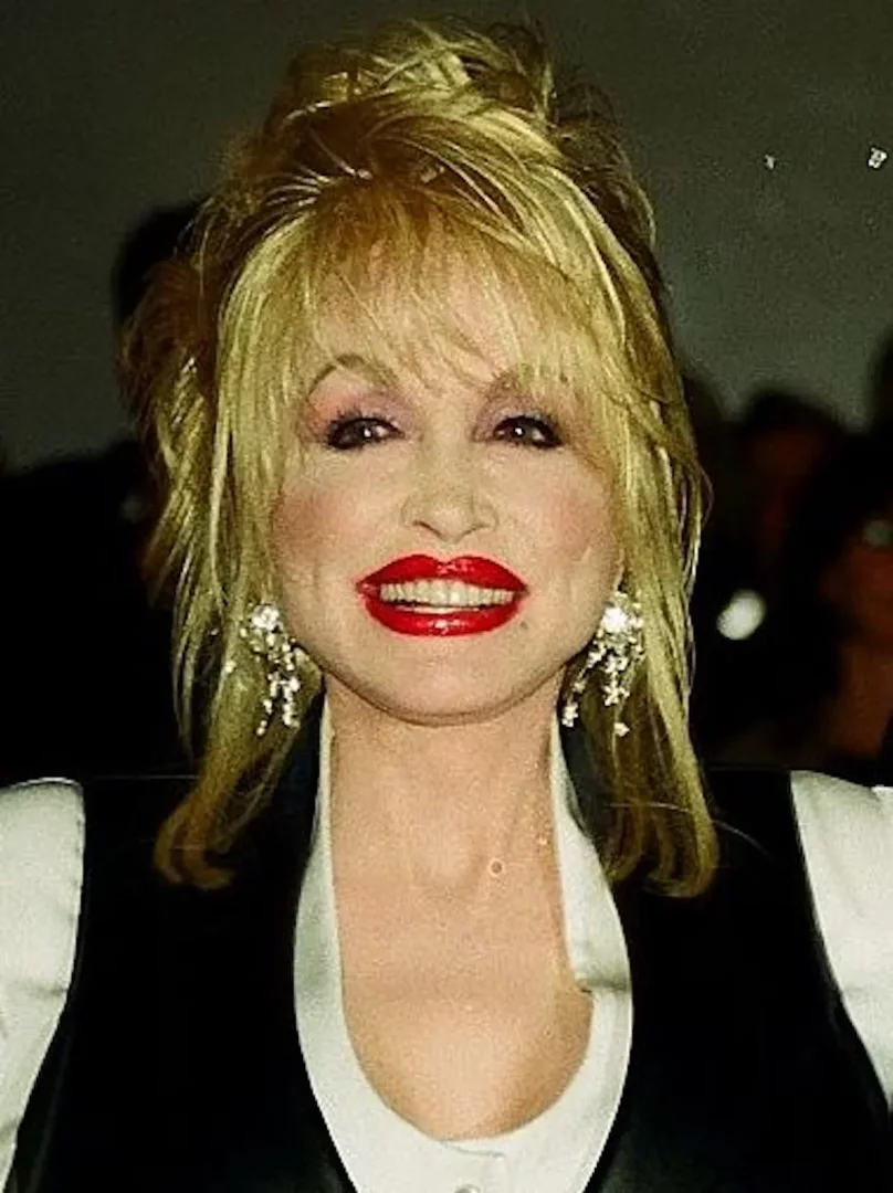 Dolly Parton'ın sarı, hacimli saçları, parlak kırmızı ruju, göz alıcı küpeleri ve samimi gülümsemesiyle yakın çekim bir portresi, onun ikonik stilini vurgulayarak sanatçının neşeli kişiliğini yansıtıyor.