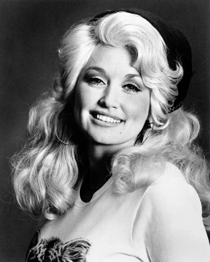 1970'lere ait bu RCA basın fotoğrafında, ikonik gür, dalgalı sarı saçları ve neşeli gülümsemesiyle kameraya bakan Dolly Parton, başının arkasında koyu renk bir bere, büyük halka küpeler ve omuzunda leopar desenli bir tişört giymiş halde duruyor.