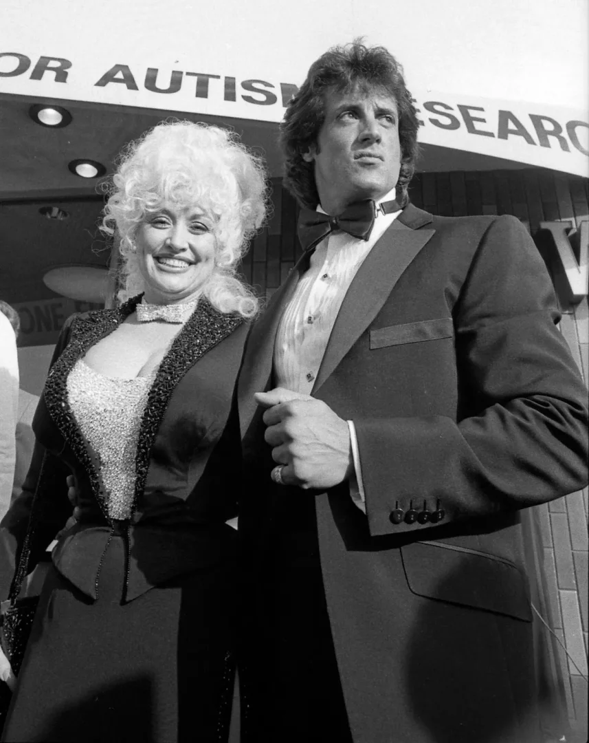 Siyah beyaz bir fotoğrafta, kıvırcık sarı saçları ve ışıltılı kıyafetiyle Dolly Parton neşeyle kameraya bakarken, siyah smokinli Sylvester Stallone ciddi bir ifadeyle ileriye doğru bakarak bir etkinlikte poz veriyor, arka planda otizm araştırmaları ile ilgili bir tabela seçilebiliyor.