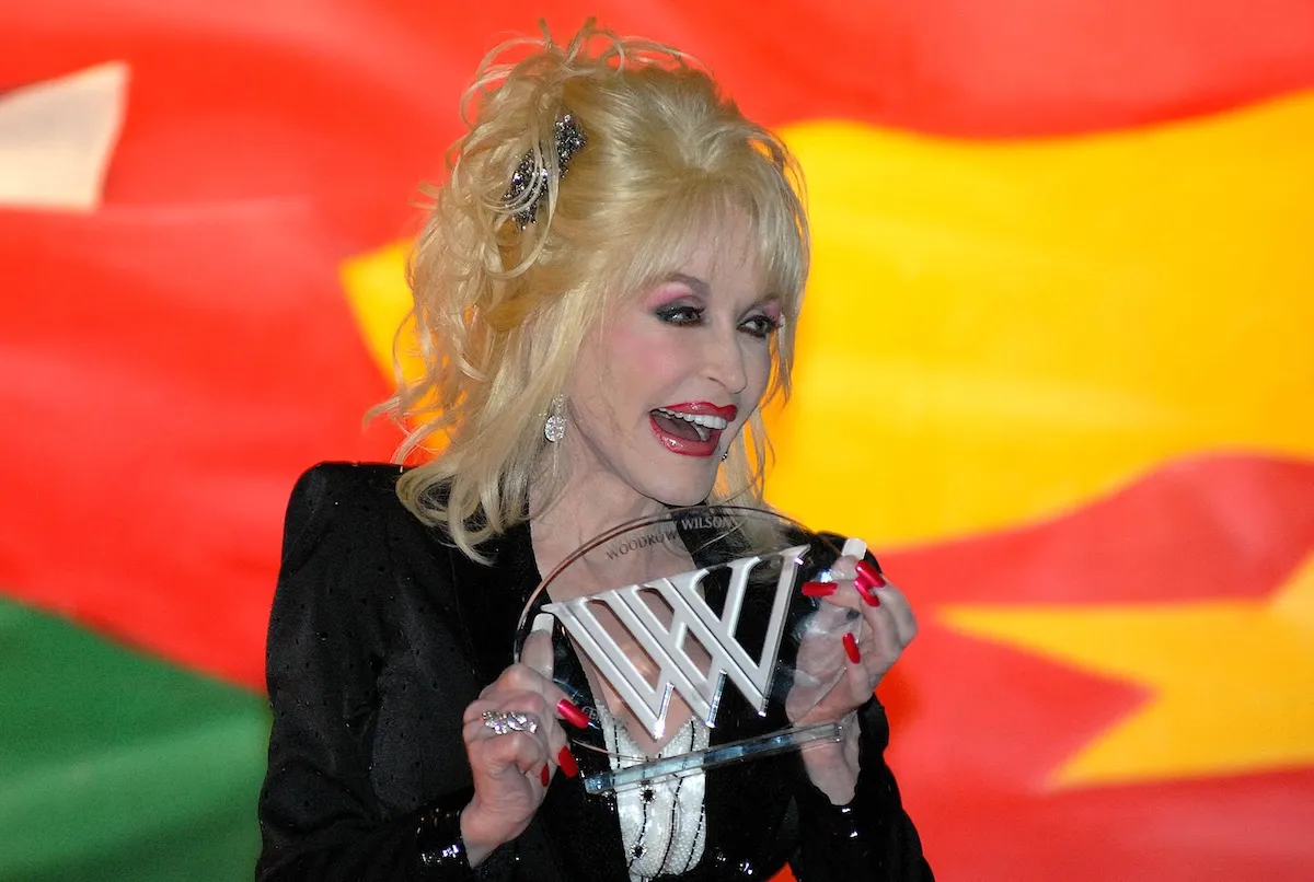 Sarı dalgalı saçları ve parlak makyajıyla dikkat çeken Dolly Parton, kırmızı ojeli tırnaklarıyla üzerinde WW ve Woodrow Wilson yazan şeffaf ödülünü gururla tutarken ağzı açık şekilde gülümsüyor, arkasında canlı kırmızı ve sarı renkli bir fon bulunuyor.
