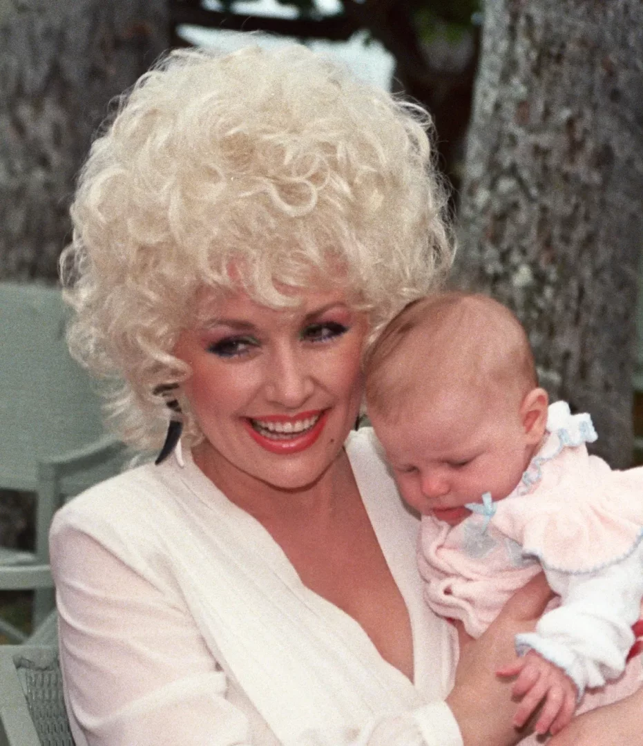 Dolly Parton, gür sarı kıvırcık saçları, parlak kırmızı ruju ve dikkat çekici makyajıyla neşeyle gülümserken kollarında uyuyan sevimli bir bebeği tutuyor ve annelik sıcaklığı yayan bu karede dış mekan bir ağacın önünde poz veriyor.