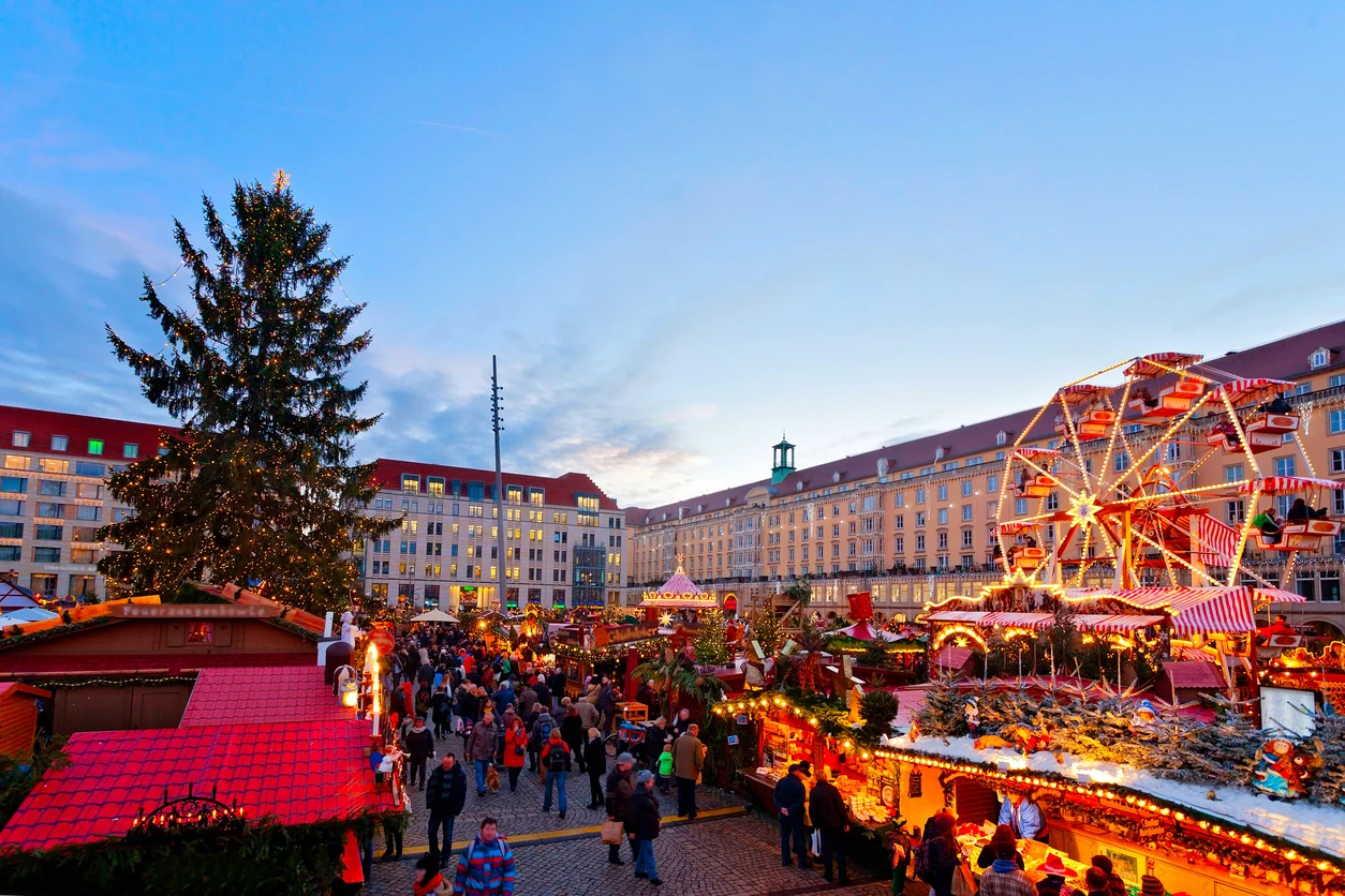 Dresden'deki kalabalık bir Noel pazarının akşam manzarası; merkezde büyük, ışıklarla süslü bir Noel ağacı, kırmızı çatılı sayısız ahşap tezgah, ışıklı bir dönme dolap ve neşeli ziyaretçilerle dolu bir alan, arkada geleneksel mimariye sahip tarihi binalarla şenlikli ve canlı bir atmosfer sunuyor.