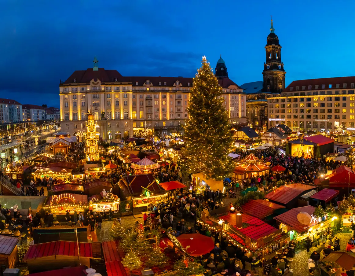 Dresden Striezelmarkt’ta ışıklarla süslenmiş Noel ağacı, ahşap stantlar ve ziyaretçiler.