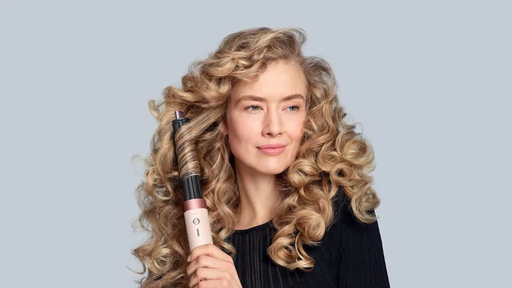 Açık gri bir fonda, altın sarısı, hacimli bukleleri olan bir kadın, saçının bir tutamını Dyson Airwrap saç şekillendiricinin Coanda hava akımıyla etrafına sararak şekillendirirken hafifçe sağa bakıyor.