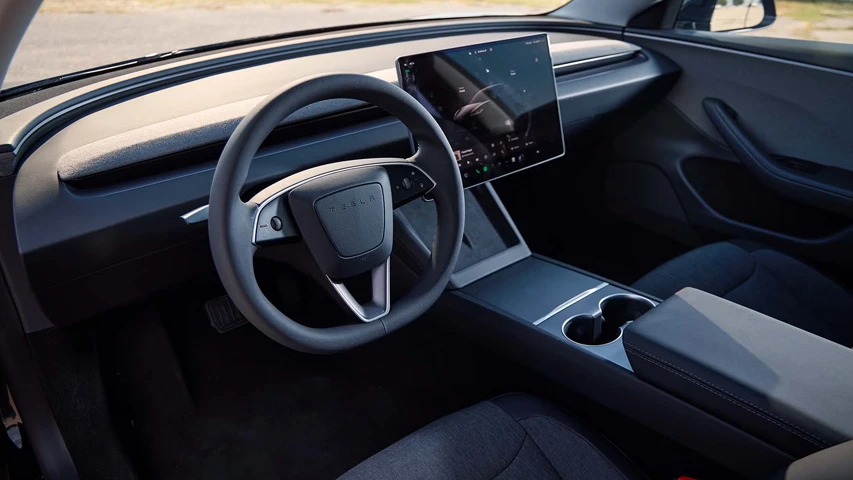 Bu Tesla elektrikli premium sedan otomobilin modern iç mekanında, sürücü koltuğundan bakıldığında ortada büyük bir dokunmatik ekran, minimalist bir ön panel ve üzerinde Tesla logosu bulunan direksiyon simidi açıkça görülüyor.