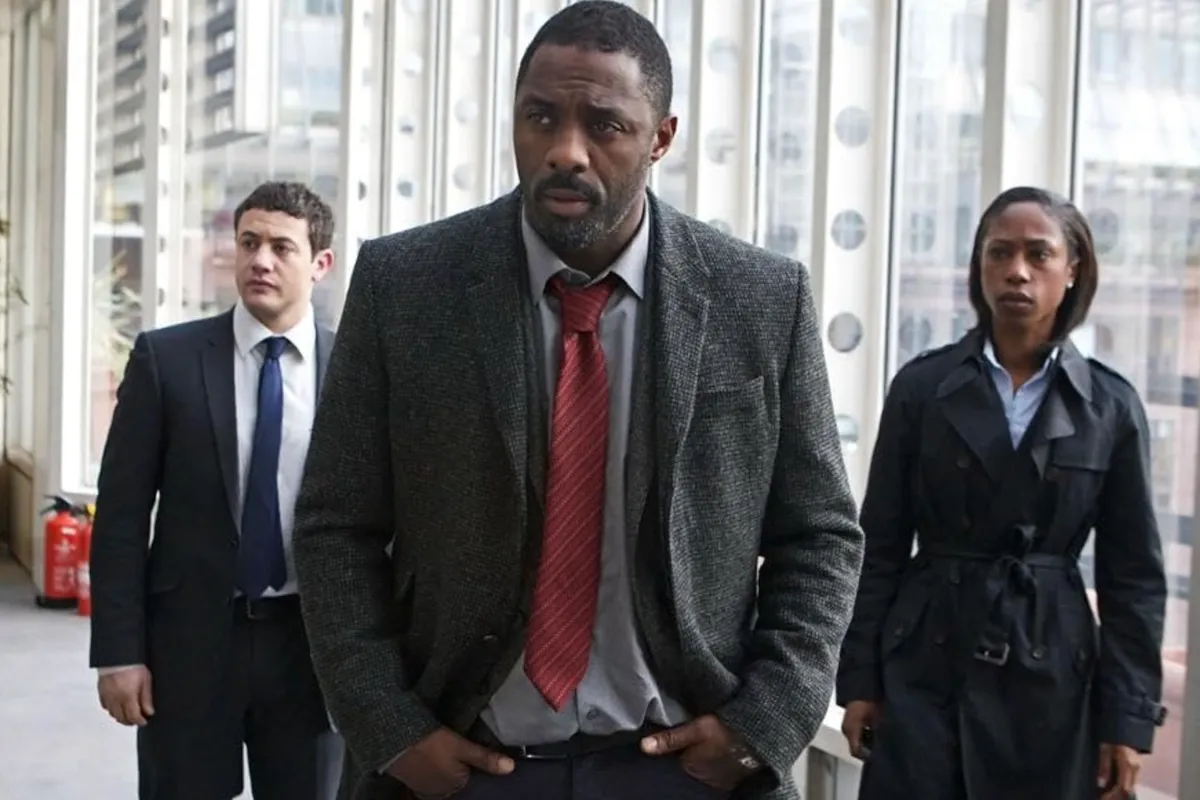 Idris Elba, Luther dizisindeki dedektif John Luther olarak koyu gri ceket ve kırmızı kravatla önde yürürken arkasında takım elbiseli bir erkek ile trençkotlu bir kadın ona eşlik ediyor; sahne en iyi suç ve polisiye dizileri seçkisinde yer alan Luther’ın karanlık atmosferini yansıtıyor.