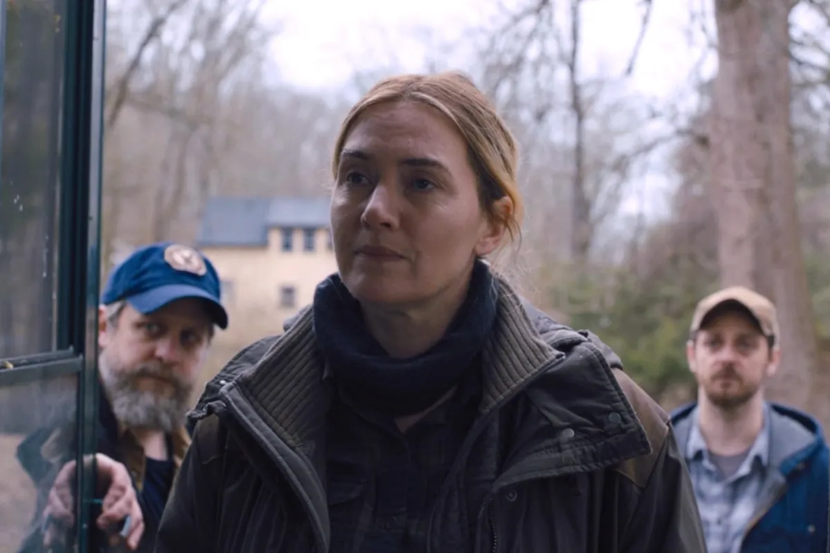 Mare of Easttown dizisinden bir sahnede, Kate Winslet’in canlandırdığı dedektif Mare Sheehan ciddi bir ifadeyle ön planda dururken arka planda iki erkek ve bulutlu havadaki ağaçlık alan görülüyor; görüntü yapımın kasvetli atmosferini yansıtıyor.