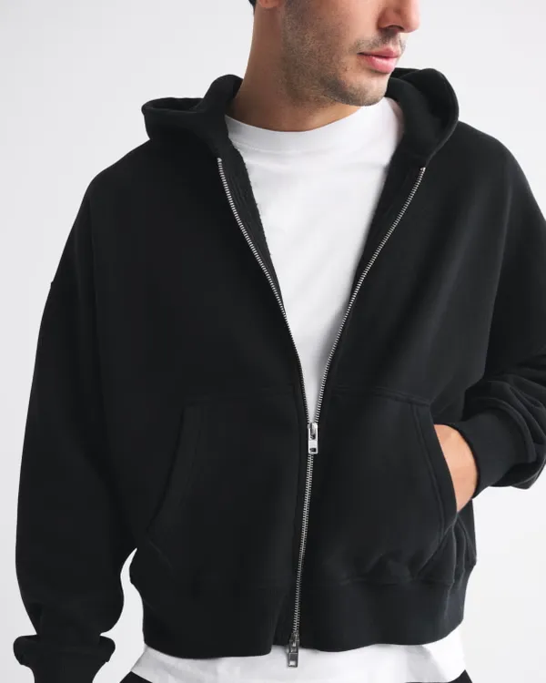 Modern ve şık bir görünüm sergileyen genç bir erkek, beyaz yüksek yakalı tişörtünün üzerine giydiği, gümüş fermuarlı, bol kesim siyah kapüşonlu erkek sweatshirt ile poz veriyor, eli cebinde.