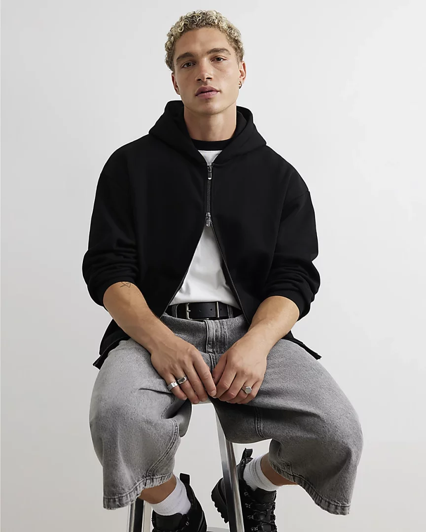 Beyaz arka planda bir taburede oturan, kıvırcık sarı saçlı genç bir erkek, siyah fermuarlı kapüşonlu sweatshirt'ünü beyaz tişörtünün üzerine giymiş, açık gri bol paça pantolonu ve siyah botlarıyla modern bir erkek sweatshirt kombinini sergiliyor.
