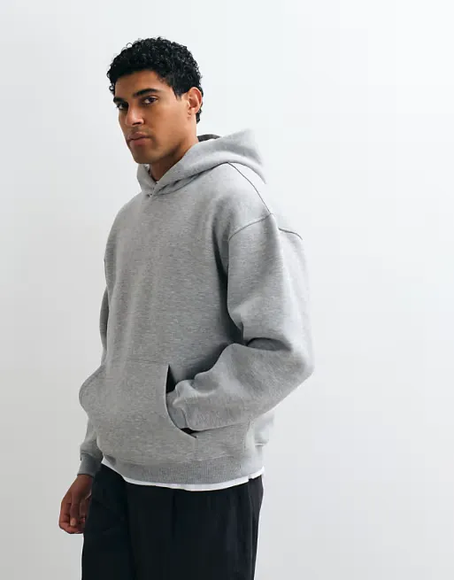 Genç bir adam, sade beyaz bir arka plan önünde, elleri geniş kanguru cebinde olacak şekilde poz verirken, açık gri renkte, modern ve rahat bir oversize erkek sweatshirt giyerek bakışlarını kameraya yöneltiyor.