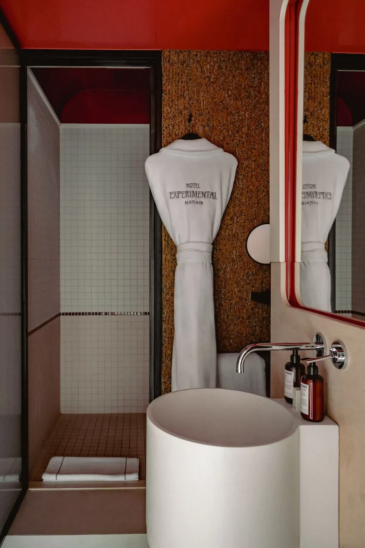 Beş yıldızlı Experimental Marais otelinin Tristan Auer imzalı modern tasarımını yansıtan bu banyoda, kırmızı tavanlı duşakabinin yanında Hotel Experimental Marais logolu beyaz bornoz, mantar görünümlü duvar ve kırmızı çerçeveli aynalı şık bir lavabo seti yer alıyor.