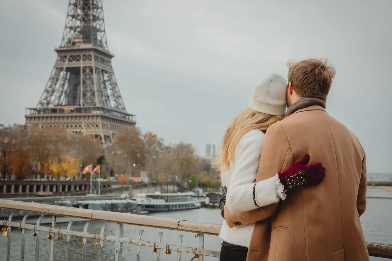 Paris'in bulutlu bir kış gününde, Eyfel Kulesi manzaralı bir köprüde, kilitlerle süslenmiş tırabzanın önünde birbirine sarılmış genç bir çift, romantik bir anı yaşarken arkadan görüntüleniyor.