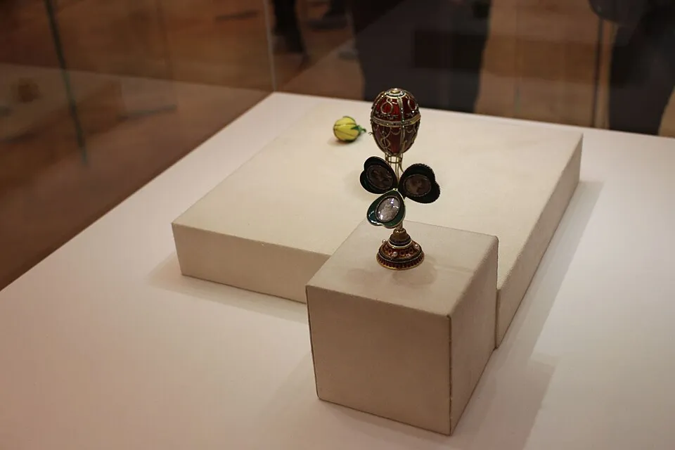 Bir müze vitrininde, altın ve kırmızı emayeyle süslenmiş, üç yapraklı yonca şeklindeki bir standın üzerinde duran ve her yaprağında portreler barındıran göz alıcı bir Faberge kraliyet yumurtası eseri ile arka planda sarı-yeşil renkli küçük bir obje, beyaz teşhir kaideleri üzerinde özenle sergileniyor.