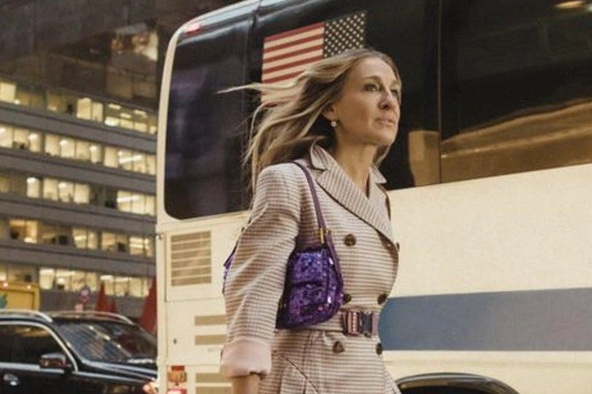 Rüzgarda hafifçe dalgalanan saçları ve çizgili trençkotuyla şehirde yürüyen Sarah Jessica Parker, parlak mor payetli Fendi Baget çantasını omzunda taşıyarak düşünceli bir ifadeyle sağa bakıyor; arka planda bir otobüs ve binalar seçiliyor.