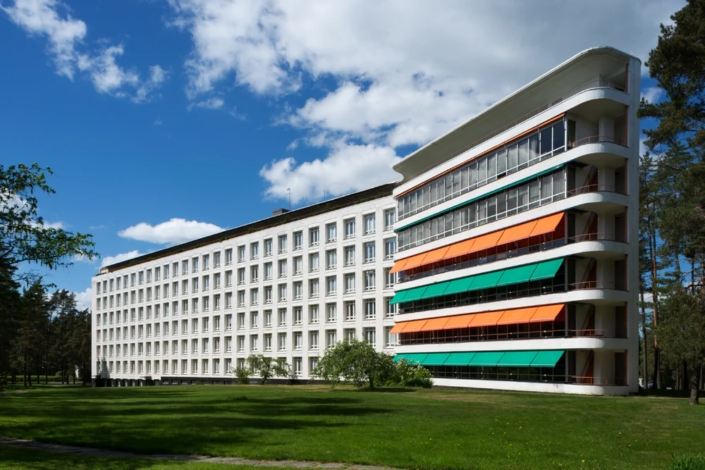 Mavi gökyüzü altında, yemyeşil bir alanda konumlanan, Alvar Aalto'nun insanı merkeze alan modernist mimari anlayışını yansıtan uzun, beyaz, çok katlı bir yapının sağ tarafında, turuncu ve yeşil tentelerin ritmik olarak sıralandığı kavisli balkonlar görülüyor.