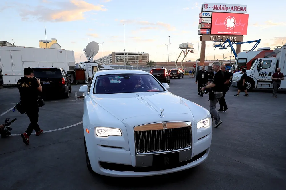 Beyaz Rolls-Royce marka lüks bir otomobil, T-Mobile Arena tabelasının ve çeşitli yayın araçlarının görüldüğü, kameramanlar, güvenlik görevlileri ve diğer kişilerin hareket halinde olduğu, muhtemelen Floyd Mayweather ile ilişkilendirilen bir etkinliğin kalabalık otopark alanında duruyor.
