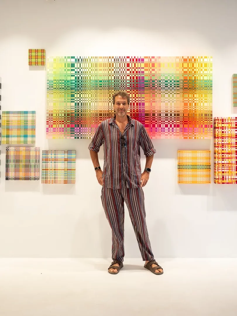 Beyaz bir duvarın önünde duran, kırmızı, mavi ve krem rengi dikey çizgili bir gömlek ve pantolon giyen, kumral saçlı ve sakallı Francesco Maccapani Missoni, arkasında dokuma kumaş desenlerini anımsatan, çok renkli küçük karelerden oluşan büyük bir sanat eseri ve çevresindeki küçük tablolarla bir sergi alanında poz veriyor.
