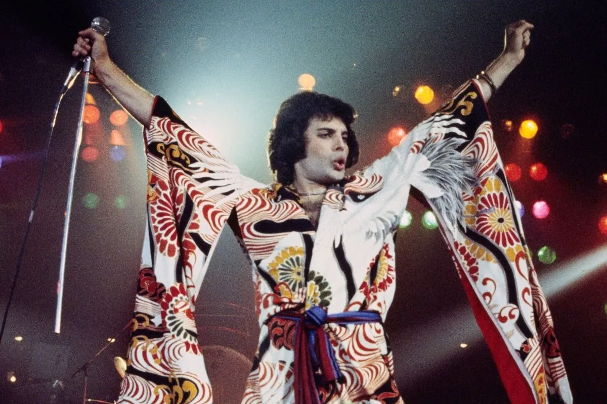 Sahne kimliğine yön veren Freddie Mercury, kolları yukarıda, mikrofonu tutarken, beyaz zemin üzerine kırmızı, sarı ve siyah desenli, kuş motifli gösterişli bir sahne kostümüyle enerjik bir performans sergiliyor, bu benzersiz kostümleri Splendid Things sergisinde de yer alıyor.