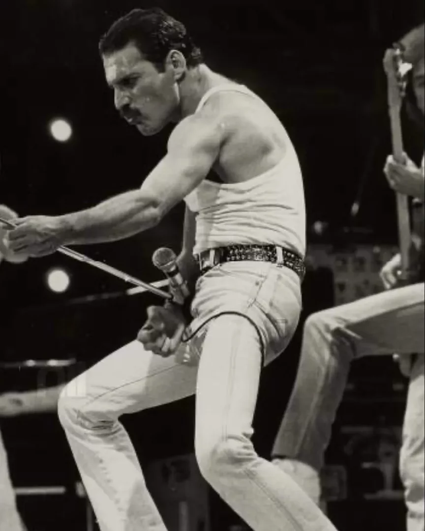Freddie Mercury, 1985 Live Aid konserinde ikonik dinamik duruşuyla, terli ve kaslı koluyla mikrofon standını ileri uzatmış, beyaz atleti, kot pantolonu ve zımbalı kemeriyle sahnede yoğun bir performans sergilerken görünüyor.