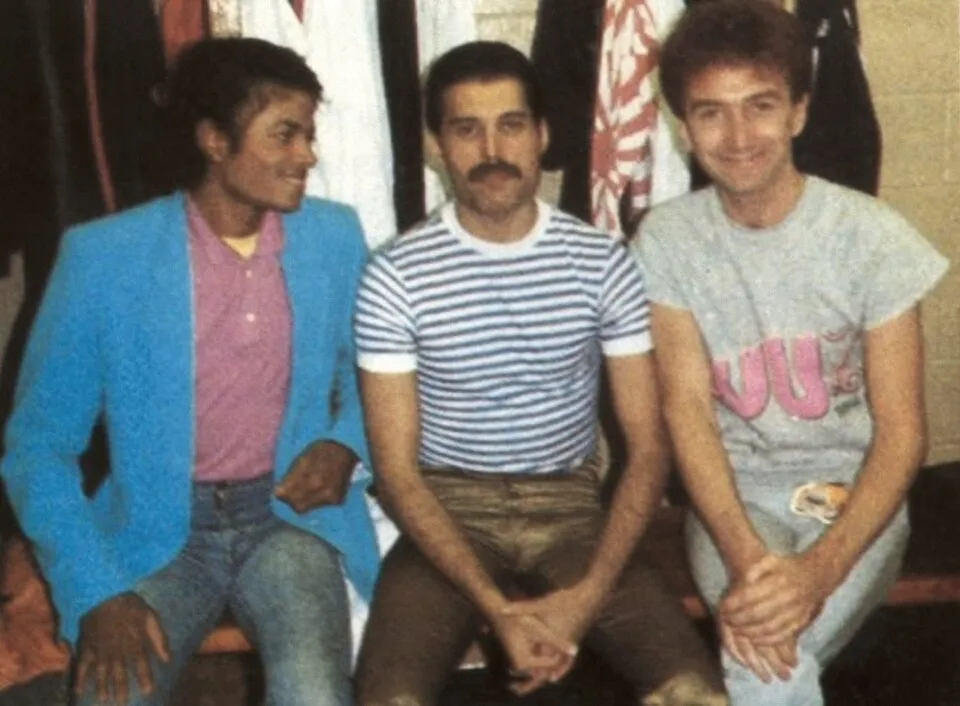 Michael Jackson mavi ceketi ve pembe polo tişörtüyle hafifçe yana dönmüş gülümserken, bıyıklı Freddie Mercury çizgili tişörtüyle düz kameraya bakıyor ve Queen'den Brian May ise gri tişörtüyle neşeyle gülümsüyor.
