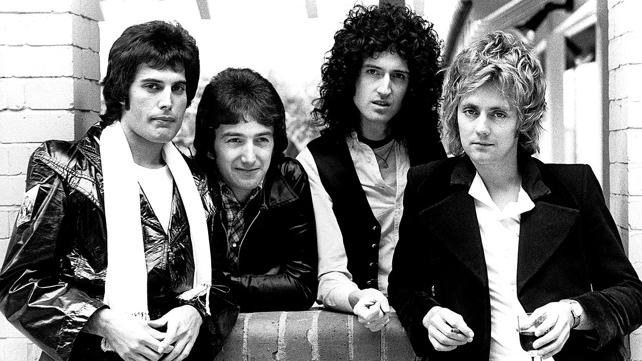 Queen grubunun kuruluş yıllarından siyah beyaz bir görselde, karizmatik duruşuyla Freddie Mercury sol başta beyaz atkısıyla, yanında John Deacon, Brian May ve Roger Taylor ile birlikte kameraya bakıyor.