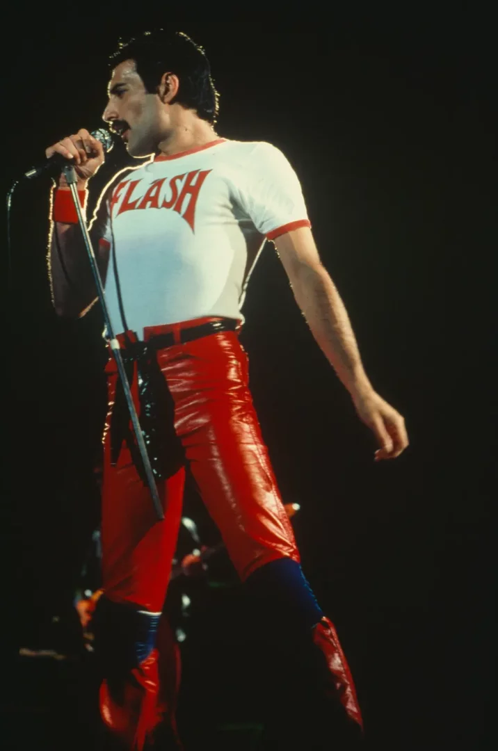 Freddie Mercury, sahnede mikrofon tutarak, üzerinde kırmızı FLASH yazısı olan beyaz tişörtü ve parlak kırmızı deri pantolonuyla şarkı söylerken görülüyor; bu görsel, bir Freddie Mercury sergisinde yer alabilecek enerjik bir anı yansıtıyor.