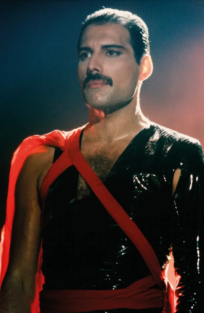 Freddie Mercury sergisi kapsamında sunulan bu çarpıcı görselde, ikonik bıyığı ve yoğun bakışlarıyla sahne enerjisini yansıtan Freddie Mercury, dramatik ışıklar altında terlemiş bir şekilde, siyah parlak bir üst, çapraz kırmızı kuşaklar ve omzundan sarkan kırmızı bir pelerinle büyüleyici bir performans anında görülüyor.