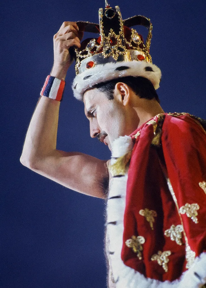 Sağ elinde tacını tutan, kırmızı ve beyaz kraliyet pelerini giymiş, bıyıklı Freddie Mercury, sahnedeki ikonik duruşlarından birinde izleyicilerine bakarken bu görsel Freddie Mercury sergisi için unutulmaz bir anı sunuyor.