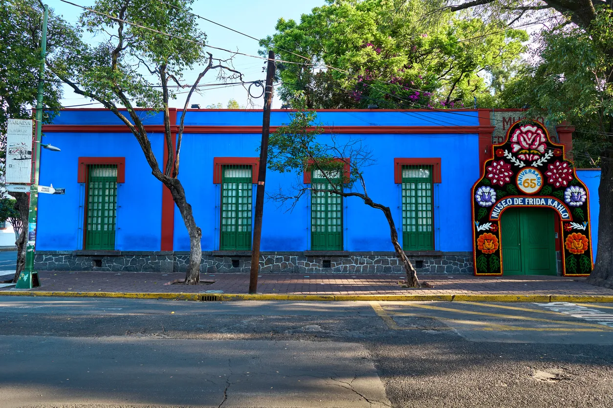 Frida Kahlo'nun Mexico City'deki Casa Azul müzesi, kobalt mavisi cephesi, kırmızı çerçeveli yeşil pencereleri, sanatçının renkli dünyasını yansıtan çiçek motifleriyle bezenmiş giriş kapısı ve önündeki ağaçlarla dolu sokak manzarasıyla parlıyor.