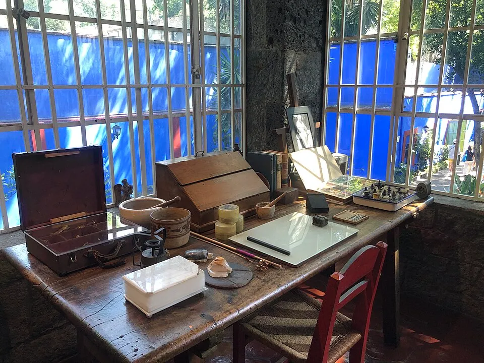 Frida Kahlo'nun Mavi Evi'nde geniş pencerelerden parlak mavi duvarların ve yeşilliklerin göründüğü aydınlık bir odada, üzerinde açık bir kahverengi kutu, havanlar, mürekkep takımı, defterler, fırçalar ve diğer kişisel eşyaların bulunduğu eski ahşap bir çalışma masası ile sağda kırmızı sandalyenin bulunduğu bir detay görülüyor.