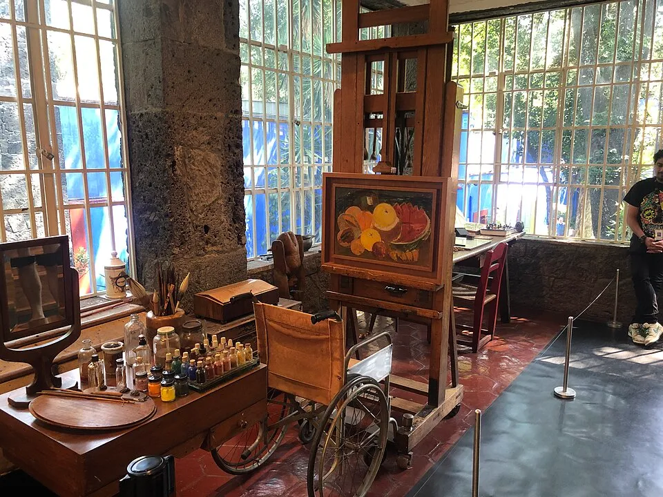 Frida Kahlo'nun Mavi Evi olarak bilinen Casa Azul'daki aydınlık stüdyosunda, tekerlekli sandalyesinin yanında duran ahşap şövalede canlı bir meyve natürmort tablosu, boya şişeleri ve fırçalarla dolu bir çalışma masası ile dışarıdaki bahçeyi gösteren geniş pencereler resmedilmiştir.