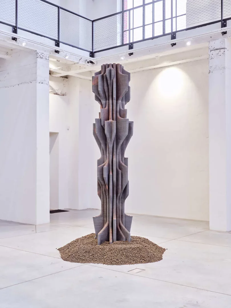Modern bir sanat galerisinin beyaz duvarları ve beton zemininde, dalgalı, katmanlı bir yapıya sahip, koyu gri, soyut bir heykel, tabanında toprağa benzer bir yığınla yükseliyor; arkasında ise asma kat ve büyük pencerelerle aydınlık bir sergi alanı görülüyor.