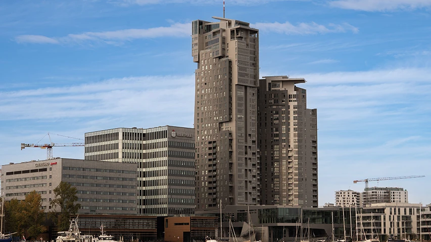 Gdynia'nın hareketli şehir merkezini gösteren bu geniş açılı görüntüde, liman boyunca sıralanmış çok katlı, modernist mimariye sahip binalar ve tekneler dikkat çekiyor; mavi gökyüzü altında uzaklarda görünen inşaat vinçleri ise yeni yapıların yükselişini vurguluyor.