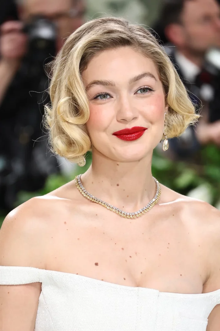 Gigi Hadid, beyaz straplez bir elbiseyle, ikonik dalgalı saç modeliyle zarif bir şekilde gülümserken, dudaklarında parlak kırmızı ruj ve boynunda ışıltılı pırlanta bir kolye ile yakın plandan görünüyor.