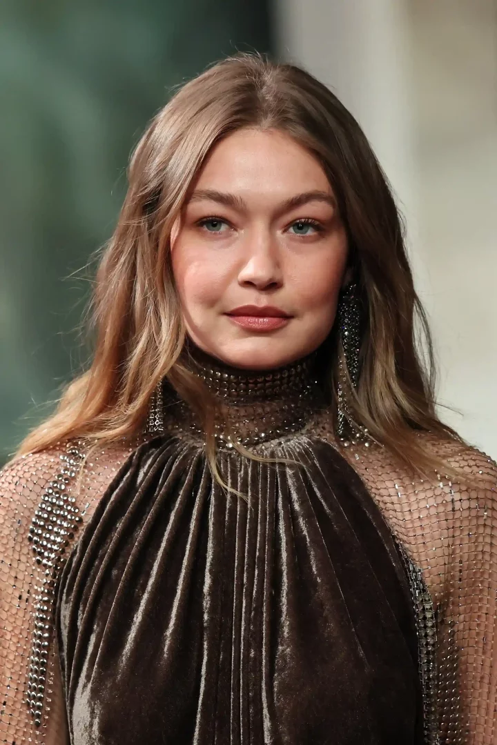 Gigi Hadid, Ralph Lauren podyumunda, ışıltılı metal file ve taş detaylarıyla süslenmiş koyu kahverengi kadife elbisesi içinde, hafif dalgalı saçları ve doğal tonlardaki makyajıyla zarif bir ifadeyle kameraya bakıyor.