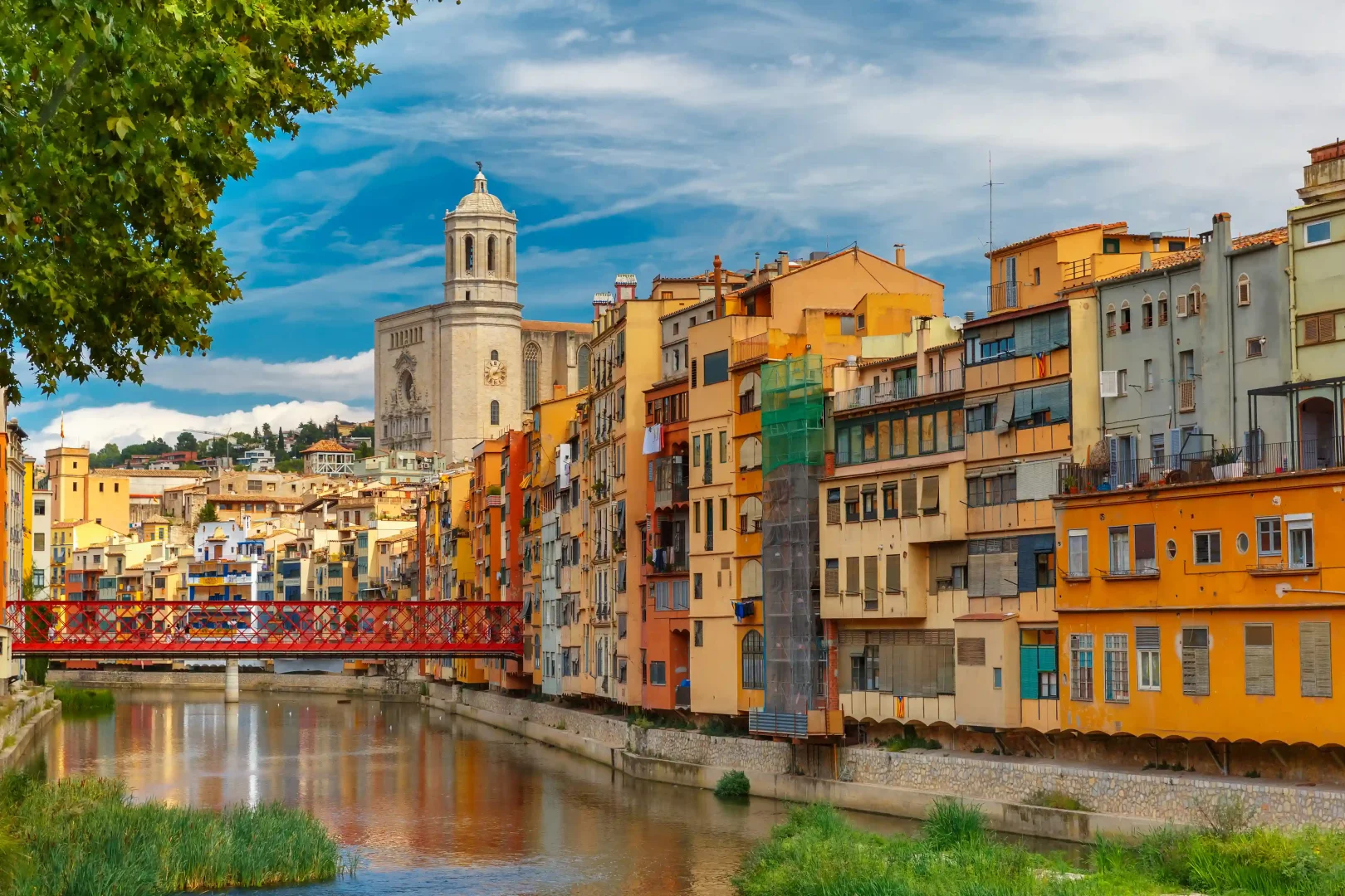 Girona'nın tarihi merkezinde, yemyeşil bitki örtüsüyle çevrili Onyar Nehri'nin iki yakasında yükselen, göz alıcı renklerdeki geleneksel evler, ikonik kırmızı köprü ve arkada heybetli Girona Katedrali'nin çan kulesiyle büyüleyici bir şehir manzarası.