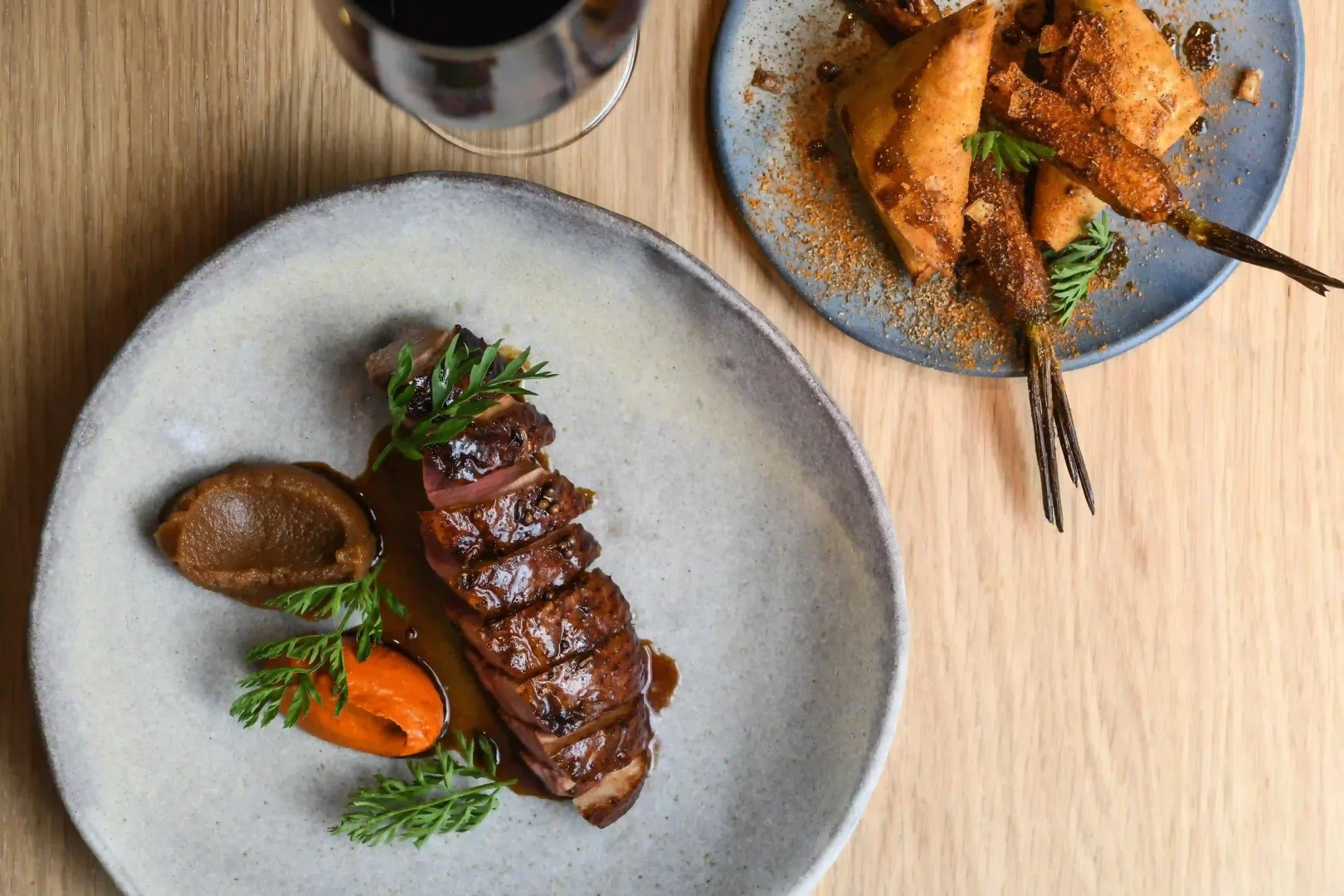Frenchie Pigalle restoranında sunulan, kızarmış derili özenle dilimlenmiş ördek göğsünün kahverengi ve turuncu püreler, taze otlarla garnitürlendiği bu karede, yanında baharatlı kızarmış havuçlar ve çıtır hamur işlerinin yer aldığı tabakla birlikte bir kadeh kırmızı şarap, Grand Pigalle Experimental'ın iyi yemek ve şarap deneyimini yansıtan bir gastronomi adresi olduğunu vurguluyor.