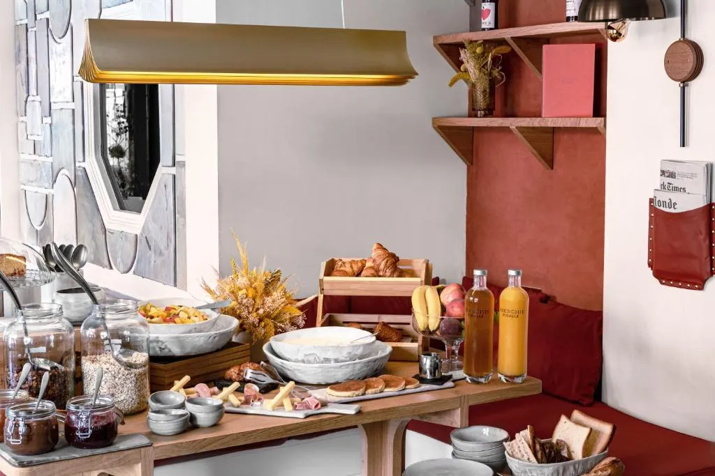 Grand Pigalle Hotel'in modern tasarımlı kahvaltı salonunda, çeşitli tahıllar, taze meyveler, kruvasanlar, pankekler, peynir ve şarküteri ürünleri sunan zengin bir açık büfe masası, tavandan sarkan şık aydınlatma armatürü ve sanatsal duvar dekorasyonuyla keyifli bir başlangıç vadediyor.