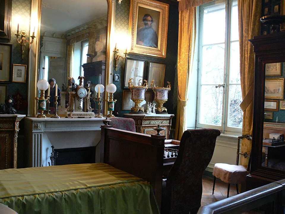 Gustave Moreau'nun Paris Evi'nin tarihi bir odasında, yeşil ve sarı çizgili örtülü bir yatak, üzerinde antika saatler ve şamdanlar bulunan mermer şömine, odanın derinliğini yansıtan gösterişli altın çerçeveli büyük bir ayna, dışarıyı gösteren desenli altın perdeli pencereler ve duvarları süsleyen sayısız tablo ve sanat eseriyle dolu, zengin ve sanatsal bir atmosfer dikkat çekiyor.
