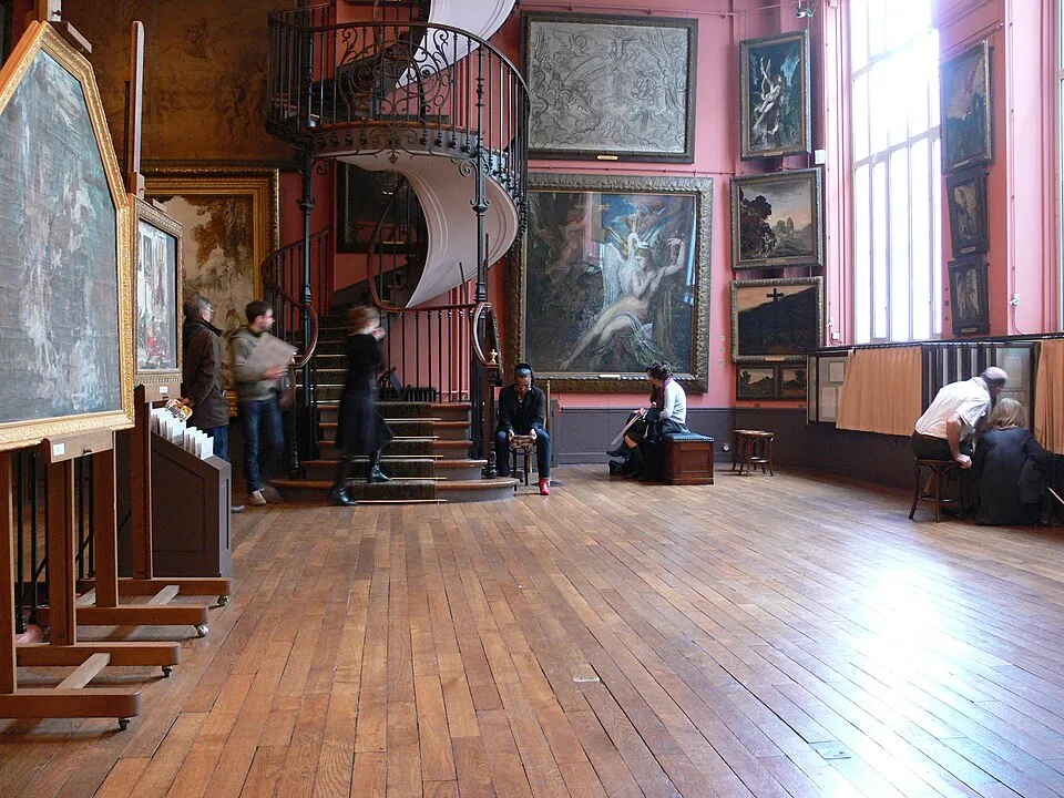 Paris'teki Gustave Moreau Evi'nin içinden, sanat eserleriyle dolu pembe duvarlar, görkemli bir spiral merdiven ve ahşap zemin üzerinde tabloları izleyen ziyaretçilerin görüldüğü, sanatsal ve tarihi atmosferi yansıtan bir görünüm.