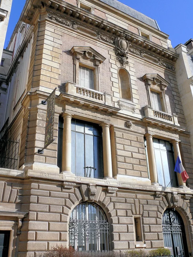 Gustave Moreau'nun Paris'teki evi olarak bilinen, klasik mimariye sahip gösterişli ve çok katlı taş bina, sağdaki penceresinden asılı Fransız bayrağı ve solunda görünen dikey pankartıyla aydınlık bir günde cepheden yukarı doğru bakışla resmedilmiştir.