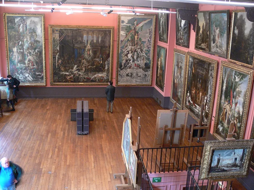 Büyük, somon rengi duvarlara sahip, ahşap parke döşemeli bir salonda, Gustav Moreau'nun resimlerine ait çok sayıda büyük ve gösterişli çerçeveli tablo duvarları baştan başa kaplarken, ziyaretçiler tabloları incelemekte ve sanatçının Paris evi ortamını deneyimlemektedir.