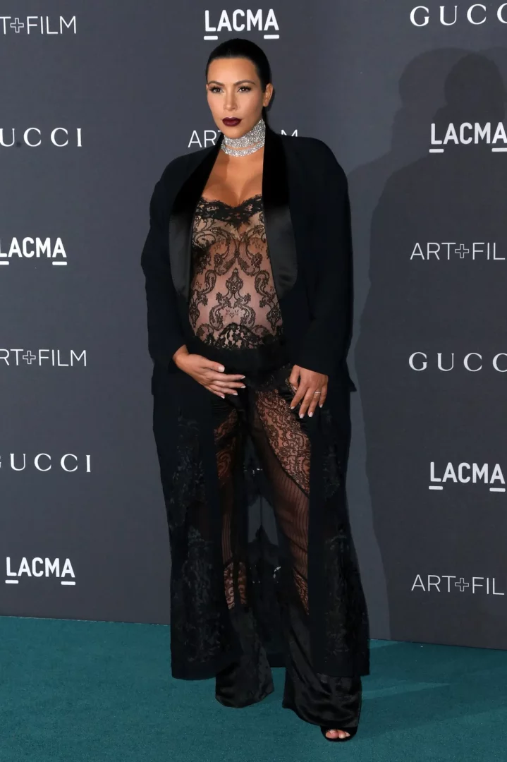 Hamile Kim Kardashian, siyah saten yakalı uzun bir ceket, parlayan bir gerdanlık, hamile karnını açıkta bırakan şeffaf dantel bir bluz ve aynı tarzda pantolonlar giyerek lacivert LACMA ve Gucci logolu bir arka plan önünde, eli karnında poz veriyor ve dikkat çekici hamile stiliyle öne çıkıyor.
