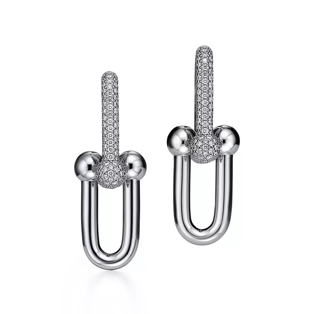 Beyaz bir arka plan üzerinde, Tiffany HardWear koleksiyonundan, üst kısımları pave pırlantalarla bezeli, ortasında pırlanta kaplı bir top ve yanlarında iki parlak metal top ile U şekilli alt kısmı birleşen şık bir çift parlak gümüş küpe.
