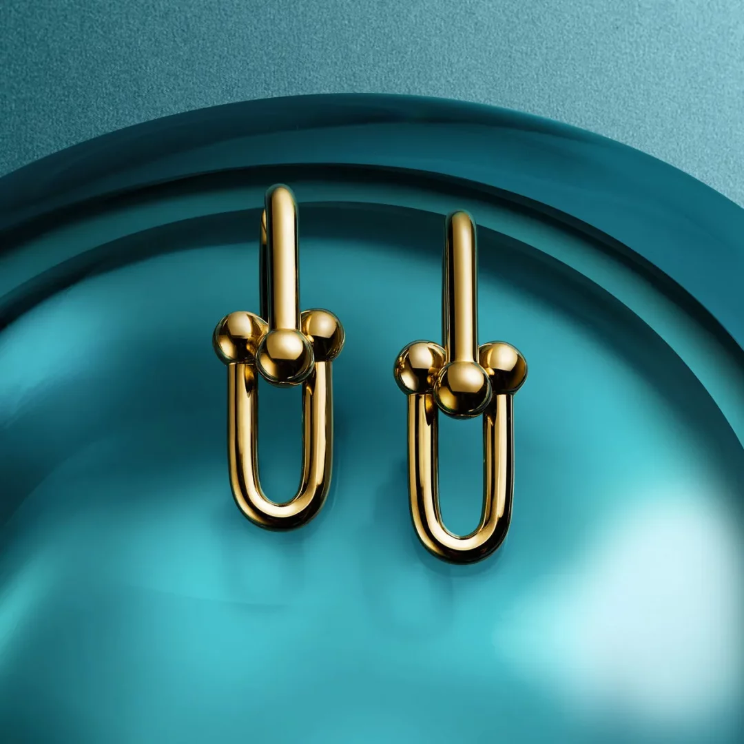 Bu görselde, parlak teal ve turkuaz tonlarında yansıtıcı bir yüzey üzerinde sergilenen, Tiffany HardWear koleksiyonuna ait olduğu anlaşılan, her biri üç küçük altın topla süslenmiş iki adet parlak altın rengi U şeklinde küpe bulunuyor.