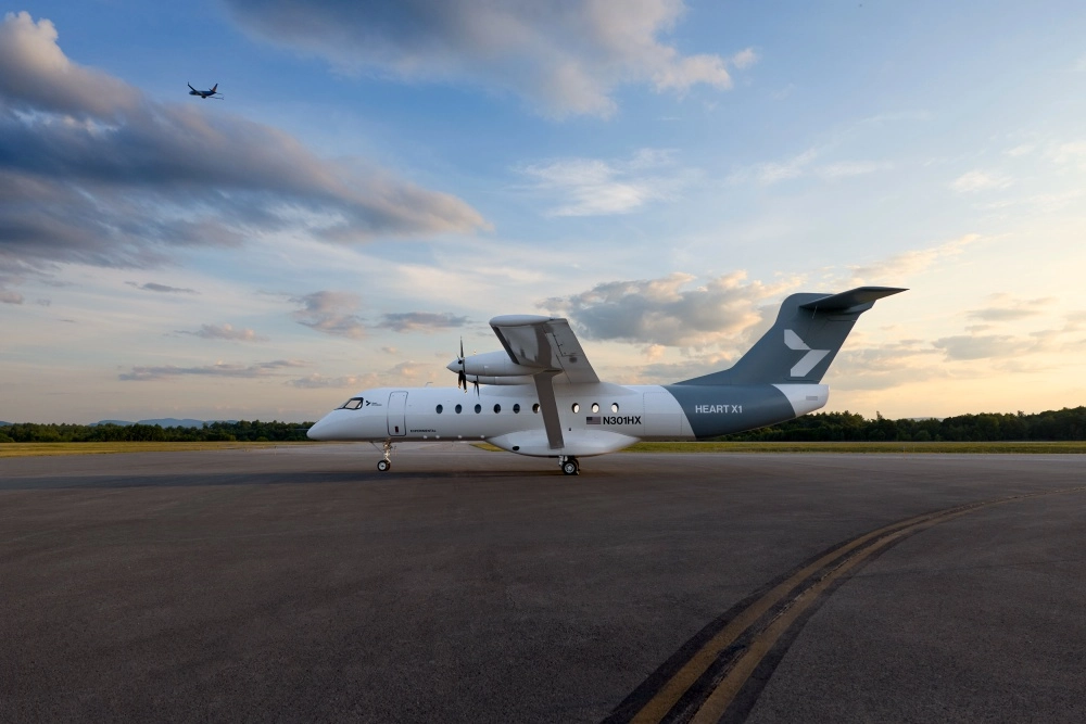 Gün batımının altın rengi ışıkları altında, pistte park halinde duran beyaz ve gri Heart Aerospace X1 elektrikli uçağı, gövdesindeki N301HX ve EXPERIMENTAL yazılarıyla dikkat çekerken, dünyanın en büyük elektrikli uçağı olarak tanıtılan bu önemli modelin üzerinde uzaklarda küçük bir uçak süzülüyor.