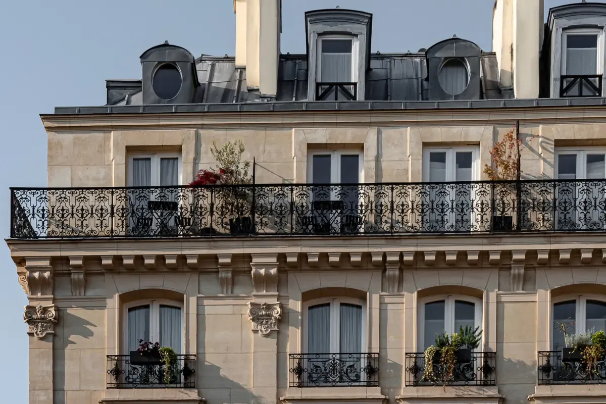 Açık renk taş cepheli, Paris tarzı bir otel binasının üst katında yer alan ferforje korkuluklu geniş balkonda zarif masa ve sandalyeler ile saksı bitkileri görülürken, çatıda estetik kavisli ve oval çatı pencereleri mimari detayları zenginleştiriyor.