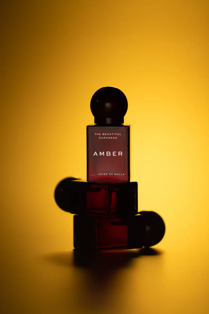 Altın sarısı ışıkla aydınlatılmış arka planda, THE BEAUTIFUL DARKNESS koleksiyonundan AMBER yazılı, koyu kehribar renkli House of Nulla parfüm şişesi, yatay yerleştirilmiş iki benzer şişenin üzerine yerleştirilmiş halde dramatik bir kompozisyon oluşturuyor.