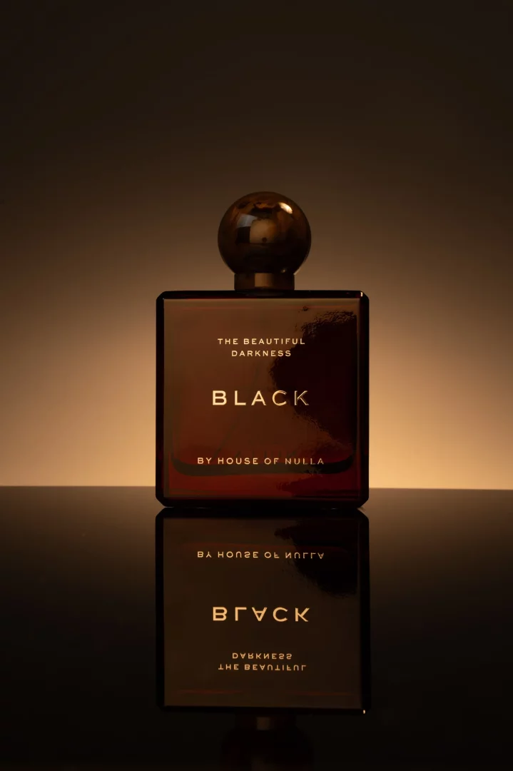 Koyu, parlak bir yüzey üzerinde duran, altın rengi yazılarla THE BEAUTIFUL DARKNESS, BLACK ve BY HOUSE OF NULLA etiketli, küre şeklinde koyu renk kapaklı dikdörtgen bir parfüm şişesi, yumuşak, sıcak bir ışıkla aydınlatılarak lüks ve gizemli bir hava katıyor.