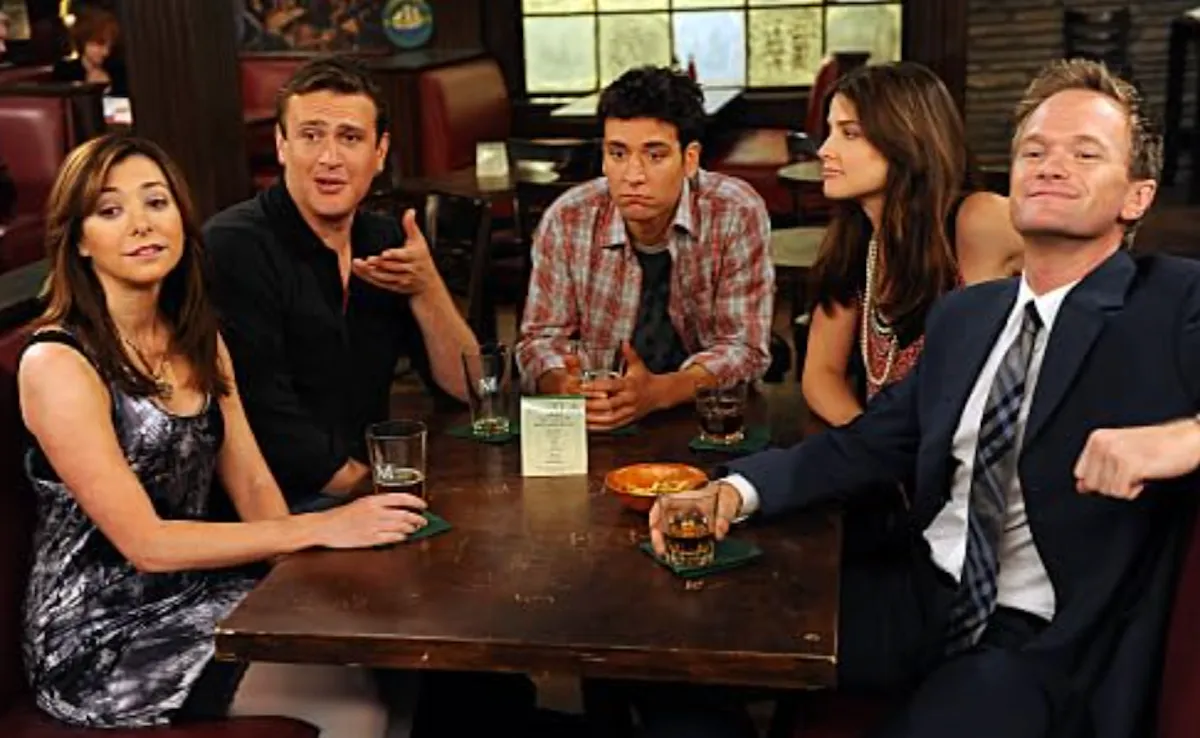 How I Met Your Mother dizisinden beş ana karakter loş ışıklı bir barda koyu kahverengi ahşap bir masanın etrafında toplanmış; Lily ve Robin dikkatle dinlerken Marshall hararetli bir şekilde konuşuyor, Ted düşünceli bir ifadeyle kadehiyle duruyor ve Barney arkasına yaslanmış, keyifli bir gülümsemeyle sohbete katılıyor, masada içkileri ve atıştırmalıklar duruyor.